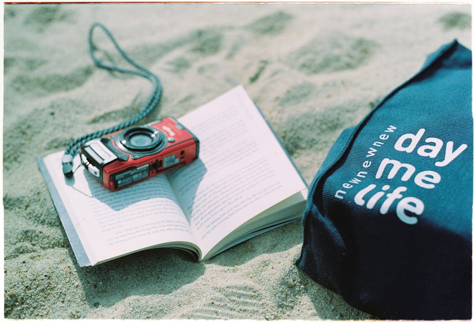 A vintage camera atop an open book next to a bag on sunlit sand, evoking a retro, carefree vibe.